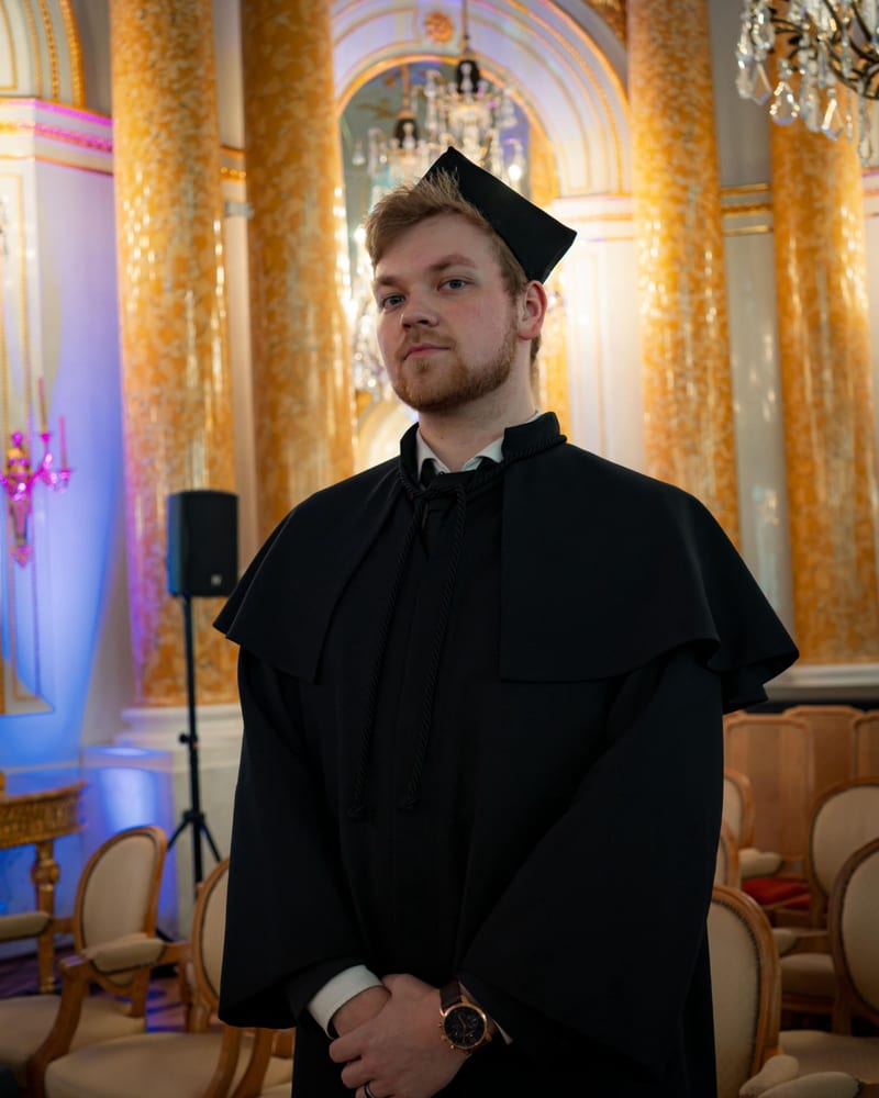 The candidate in a black academic gown and cap, standing in a gilded hall